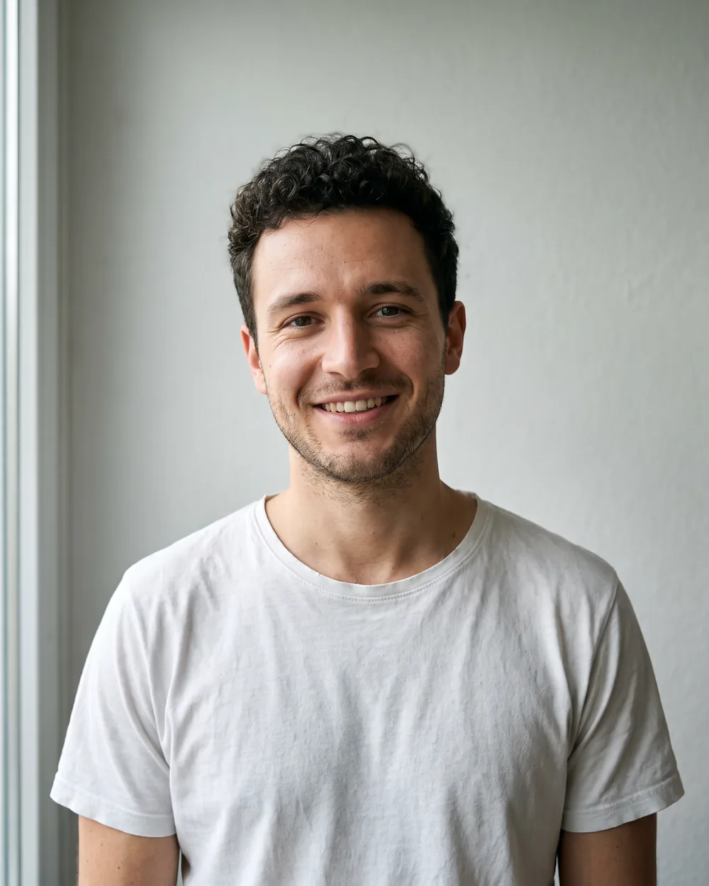 A man with curly dark hair in a white t-shirt against a grey wall