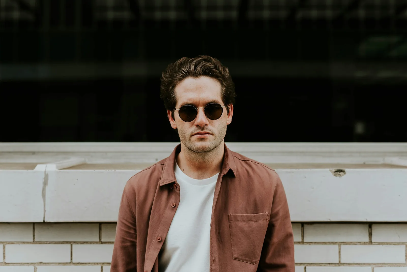 A man in sunglasses and a brown shirt against a brick wall