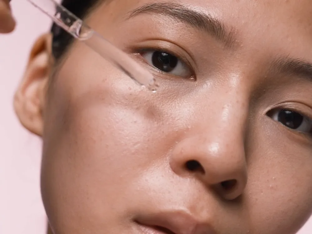 A woman holding a dropper to her cheek against pink
