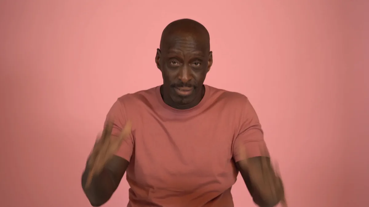 A man gesturing to camera against a pink backdrop